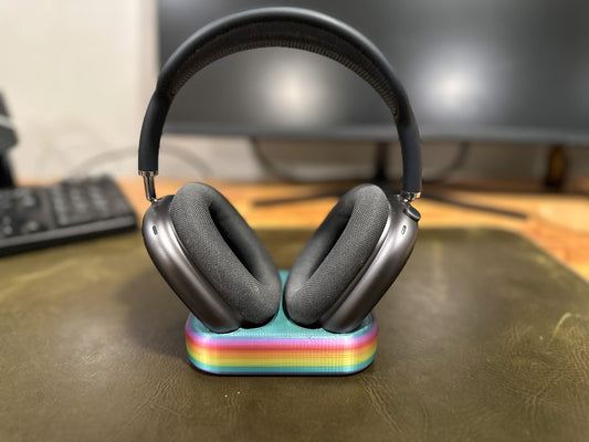 A rainbow colored head phone cradle with a pair of black airpod maxes in it all of it sitting on a desk