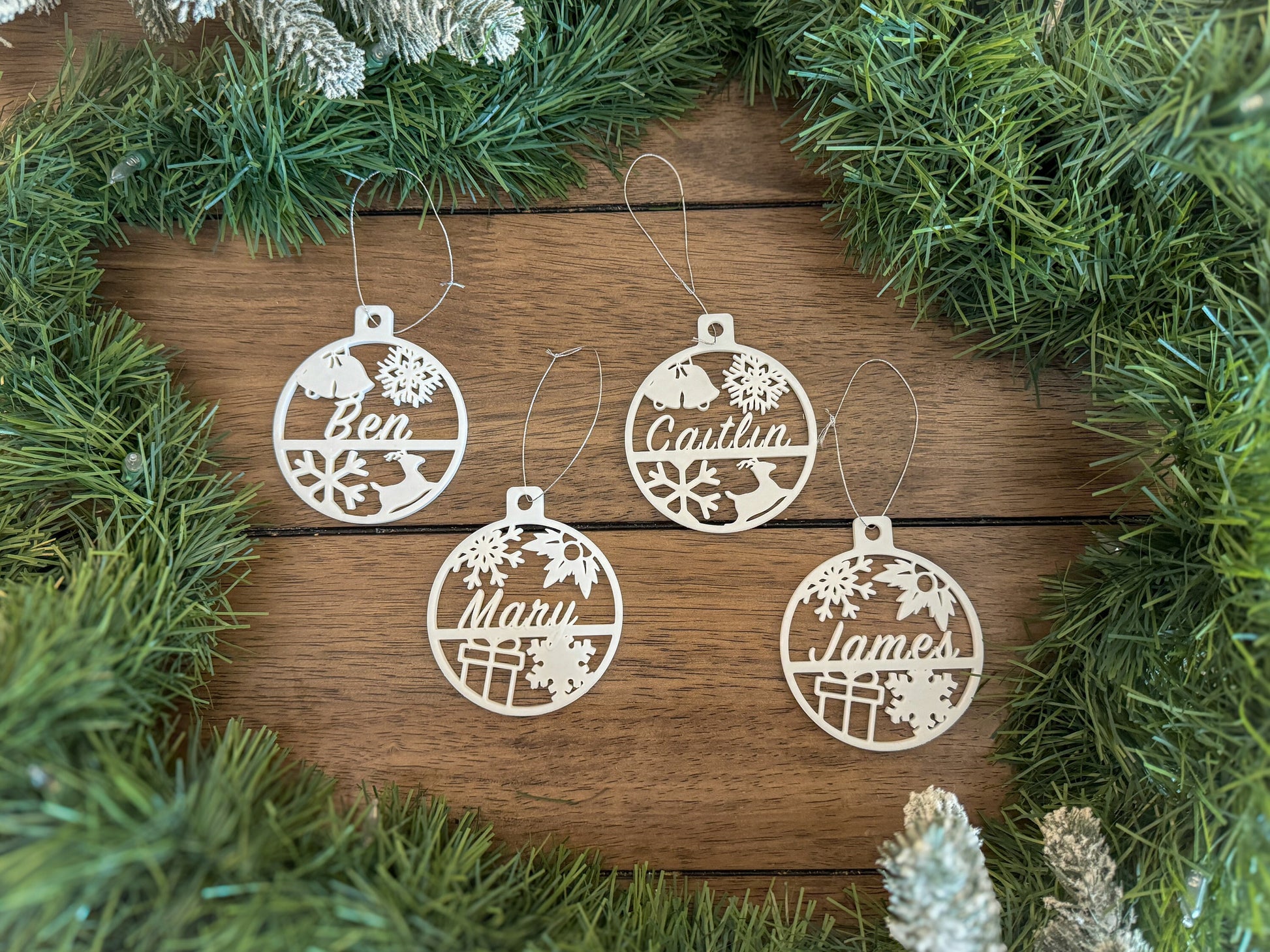 A picture of 4 circular white christmas tree ornaments with various designs of bells, snowflakes, deer, presents and holly.