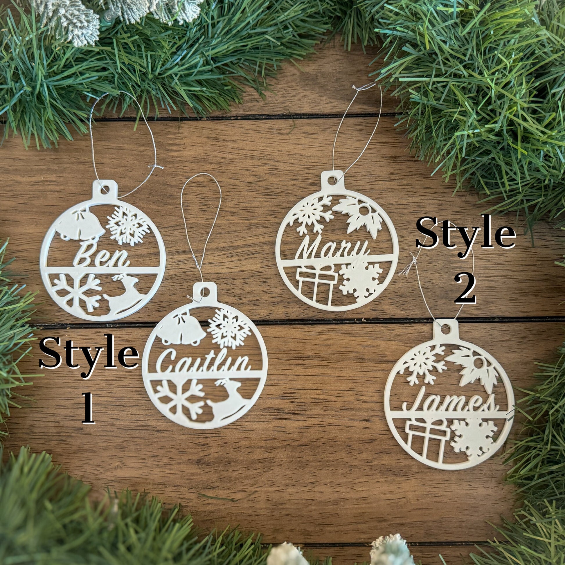 4 white circular christmas ornaments on a table showing two styles. Style one is a name, bells, snow flakes, and a deer. Style 2 is a name, snow flakes, holly, and a present.