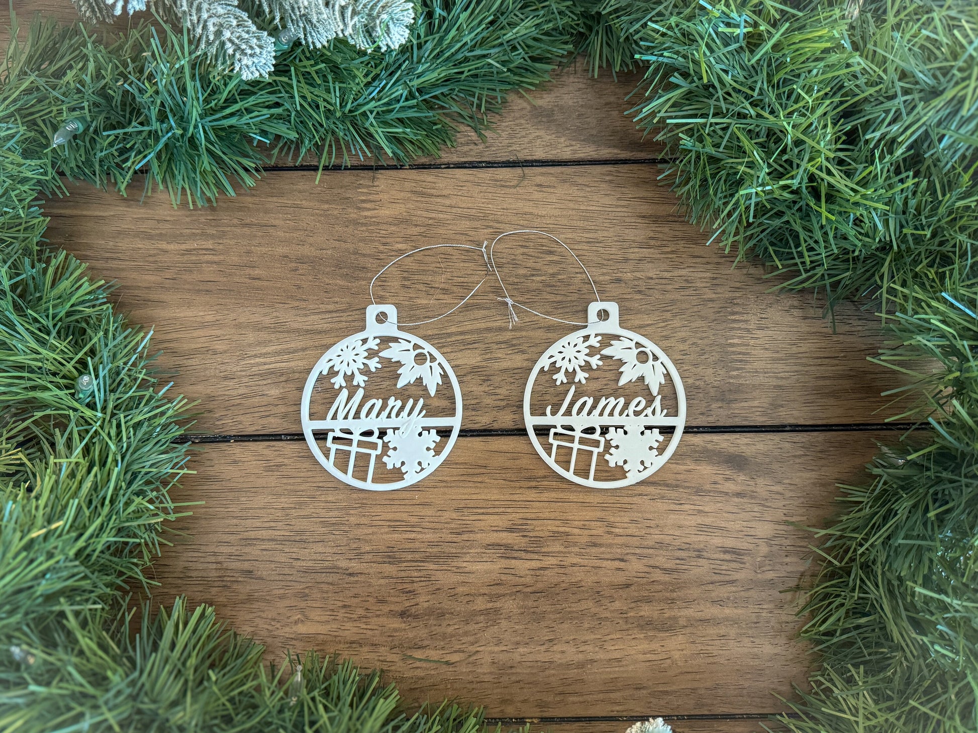 two white circular christmas tree ornaments that have a name in them with snow flakes, holly and a present