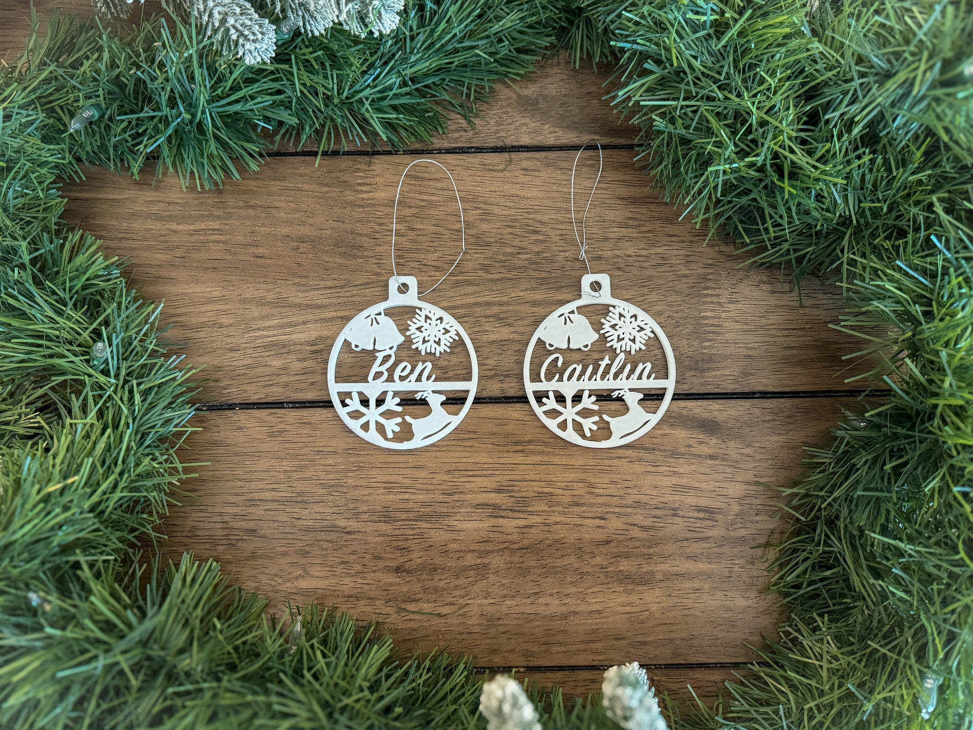 two white circular christmas tree ornaments that have a name, a bell, snow flakes and a deer.