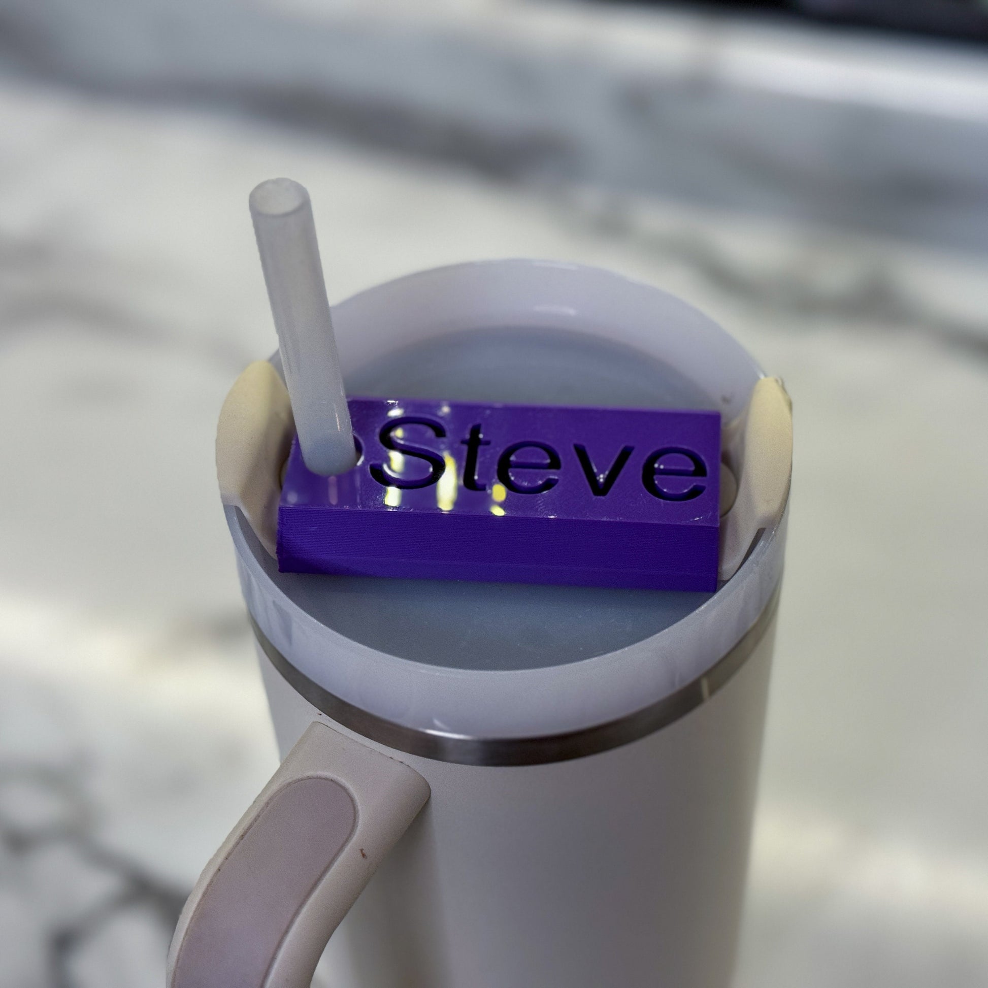 A purple name plate with black lettering that says Steve on top of a 64oz stanley tumbler