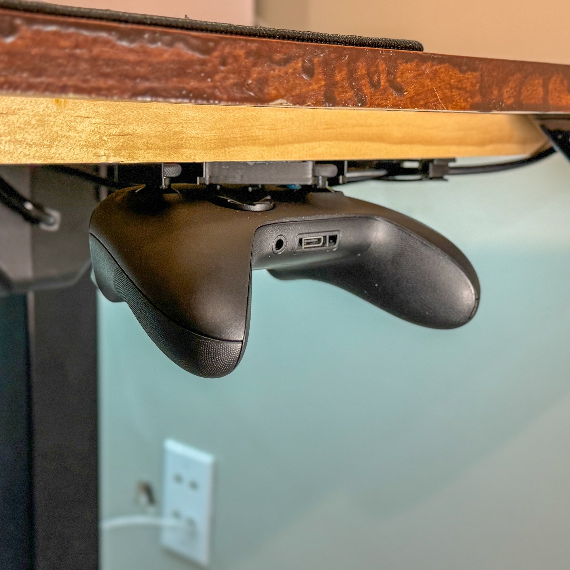 A black xbox controller hanging from an under desk mounted controller holder.